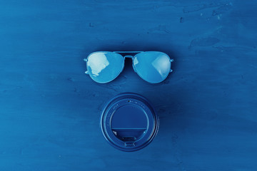 Top view of takeaway coffee cup with aviator sunglasses on classic blue background
