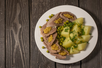 boiled beef tongue with potatoes, green peas, herbs and mustard on dark wooden background 
