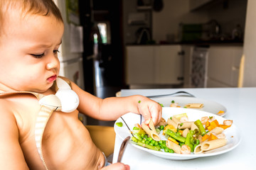 Baby sitting on his high chair eats by himself with a tasteful face.