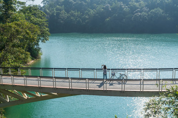 young beautiful girl stand near a bicycle while take a photo of the view in front on bike trail at the lake in the morning. Active people. Outdoors