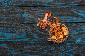 Granola with dried fruits in a glass jar and a wooden spoon on a blue wooden background.