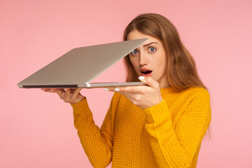 Portrait of astonished ginger girl in sweater holding half-opened laptop and looking at camera with...