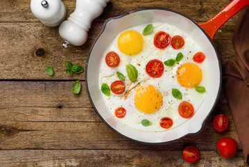 Fried eggs with cherry tomatoes and basil in a red pan for breakfast. Wood background. Top view