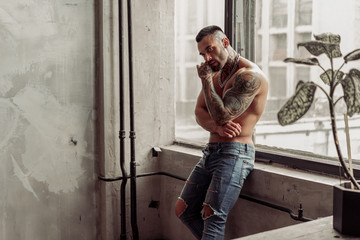 Fashion portrait of Sexy naked male model with tattoo and a black beard standing in hot pose on near the window. Loft room interior with grey concrete wall. Professional Studio image.