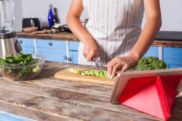 Closeup of female hands chopping cutting vegetables with knife, woman in apron cooking salad in kitchen preparing vegetarian food, following recipe on tablet, basket of vegetables and blender on table