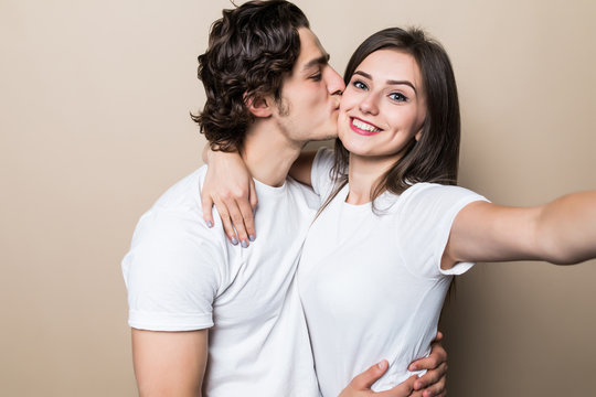 Young lovely couple posing together with klosed eyes while making selfie and man kissing woman over beige background