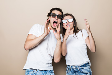 Two young shocked lovers are amazed with wide open eyes, mouthes, fixing sunglasses, in a casual wear on beige background