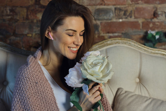 Beautiful young woman holding and sniffing big rose flower with earphone in ear on the classic coach under wool plaid with red brick wall on background at cozy home