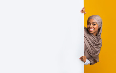 Smiling afro girl in hijab standing next to white advertisement board