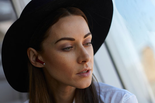 Young serious face street style fashion modern business woman in black hat and white shirt talks and listening wireless earphones in ear seating near panoramic window with city center and sky view