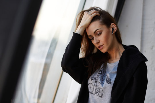 Young street fashion woman in black hoodie and grey print shirt use to talk or listen music wireless earphones in ear near panoramic window in loft interior and fixes brown hair and looks down