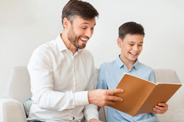 Father And Son Reading Book Sitting On Couch At Home