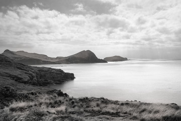 Seascape on a cloudy early morning with volcanic rocks and grass on foreground and middle ground and a rocky island and clouds in the background, Black and White Photo, Madeira Island, Portugal