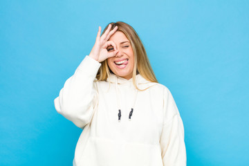 young blonde woman smiling happily with funny face, joking and looking through peephole, spying on...
