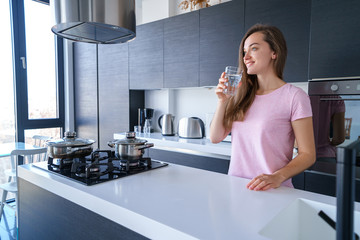 Happy attractive joyful brunette female drinking fresh clean filtered purified water at kitchen at...