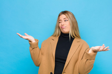 young blonde woman shrugging with a dumb, crazy, confused, puzzled expression, feeling annoyed and...