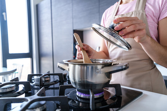Female housewife in apron using steel metal saucepan for preparing dinner in the kitchen at home. Kitchenware for cooking food