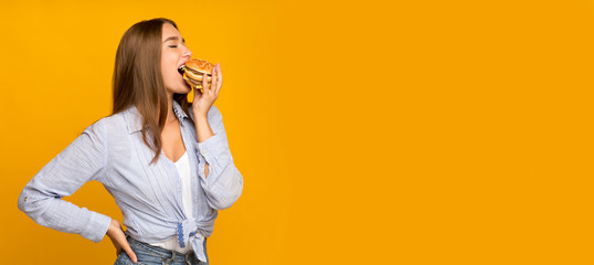 Millennial girl eating burger standing on yellow studio background, panorama