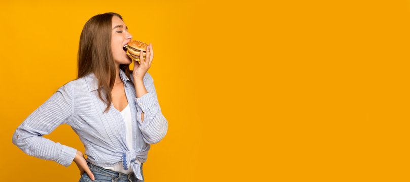 Millennial Girl Eating Burger Standing On Yellow Studio Background, Panorama