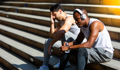Fototapeta na wymiar fitness people and lifestyle concept. African american and friend white people sweating taking a break listening to music on phone after and holding water bottle after running sitting on stair city