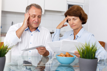 Sad mature couple at kitchen table sitting with  financial  documents