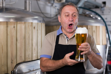 Brewer is standing with beer with foam in glass on his workplace