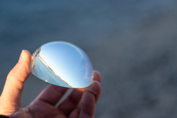 Hand holding a crystal glass hemisphere, blue reflections of surrounding sky and sea.