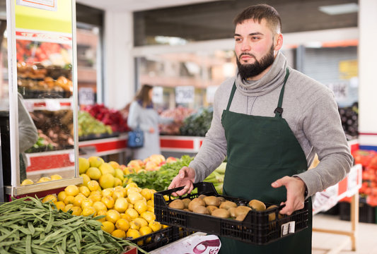 Positive man employee showing box with kiwi in grocery shop