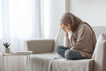Depressed arabic girl in hijab crying on sofa at home