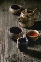 Hot black tea in different traditional wabi sabi style fireclay ceramic craft cups served with plate, teapot and bowl of dry tea leaves on old wooden table. Dark rustic atmosphere.