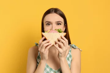 Foto auf Acrylglas Snack Junge Frau, die leckeres Sandwich auf gelbem Hintergrund isst