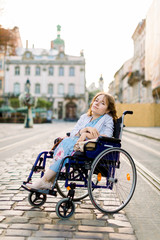 Fototapeta na wymiar A girl in blue dress in a wheelchair outdoors in the city. The disabled person enjoys life. Young beautiful disabled woman