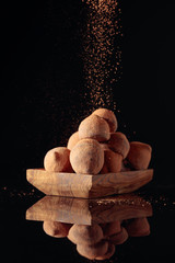 Chocolate truffles in small wooden dish sprinkled with cocoa powder. Black reflective background.
