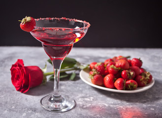 Red exotic alcoholic cocktail in clear glass, plate with fresh strawberries and red rose on the concrete background for romantic dinner.