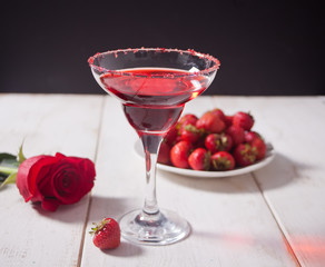 Red exotic alcoholic cocktail in clear glass, plate with fresh strawberries and red rose on the wooden white table for romantic dinner.