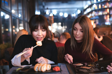 Girls eating a dinner at restaurant. Brunette is eating sushi and her girlfriend is ordering food from a menu.