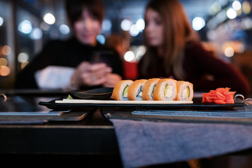 Close up shot of salmon sushi chopstick and ginger with two women watching at smartphone at the...