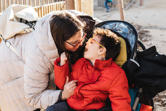 una madre juega con su hijo con pluridiscapacidad en un parque, el ni&ntilde;o va con silla de ruedas, es el invierno y van con ropa de abrigo.la mujer le saca la lengua, le hace cosquillas y rien juegan