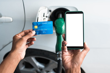 Cropped top view of man holding credit card and smartphone over gun petrol refueling at gas station.