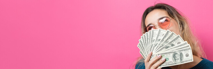 Closeup of young beautiful woman in a blue sweater in glasses with american dollars money in hand over pink background. Expresses emotions and gestures of joy.	