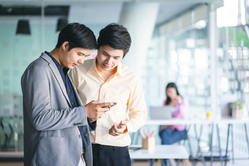 Young Asian businessmen using a smartphone and digital notebook to discuss information in a modern business working space.