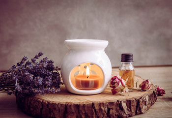 Vintage style picture of white ceramic candle aroma oil lamp with essential oil bottle and dry flower petals on natural pine wood disc, dry background with copy space. - obrazy, fototapety, plakaty