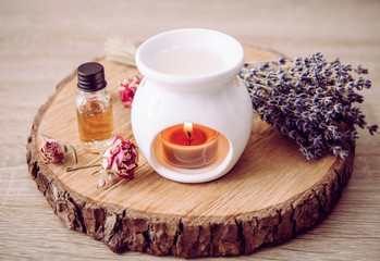 Vintage style picture of white ceramic candle aroma oil lamp with essential oil bottle and dry flower petals on natural pine wood disc, dry background with copy space.