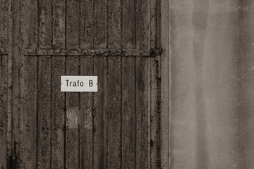 Detail of an old wooden door with metal fittings to a transformer room, old door with rusty fittings and peeling paint in detail, the sign reads: "Transformer B", black and white photo
