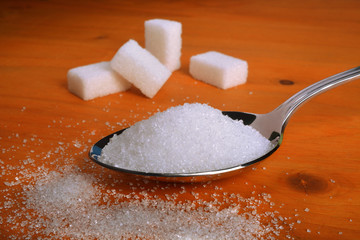 A spoon full of refined white sugar on a wooden table and sugar cubes on background..
