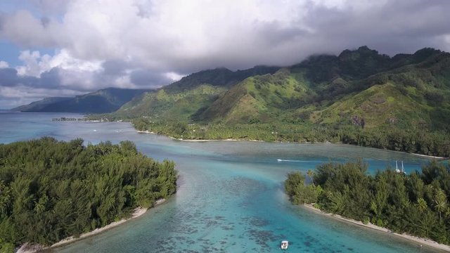 Aerial drone through the lagoon on the island of Moorea, French Polynesia. Clouds envelope the mountains in the distance as a speedboat moves through the tranquil scene.