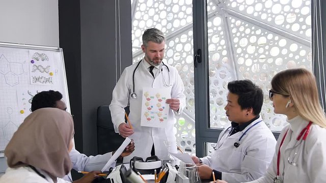 Head doctor with gray hair explaining to his considerate multiethnic doctors-colleagues report's datas with chemical compounds in boardroom