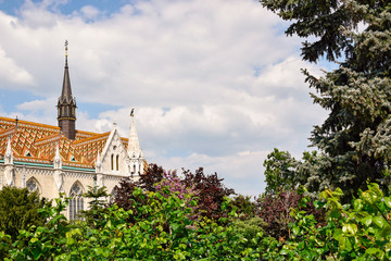 View of ornately colored tile roof of St. Matthias Church through the lush greens of trees on cloudy sky background. Fisherman's Bastion in Budapest.