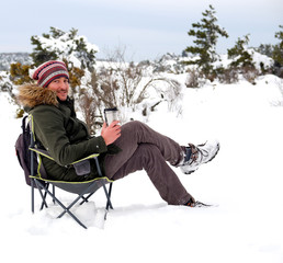Young man is sitting on camping chair, drinking coffee with thermos mug and enjoying snow in winter season.