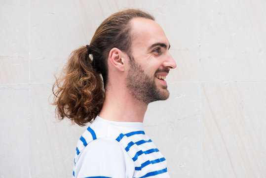 Side Portrait Of Young Man With Long Hair In Ponytail Smiling By White Background